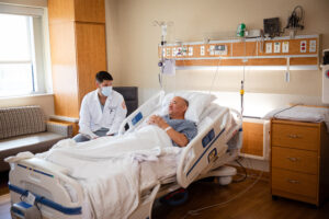 Brandon Zsigray, seated at the side of a patient bed in a hospital room, talks to a patient